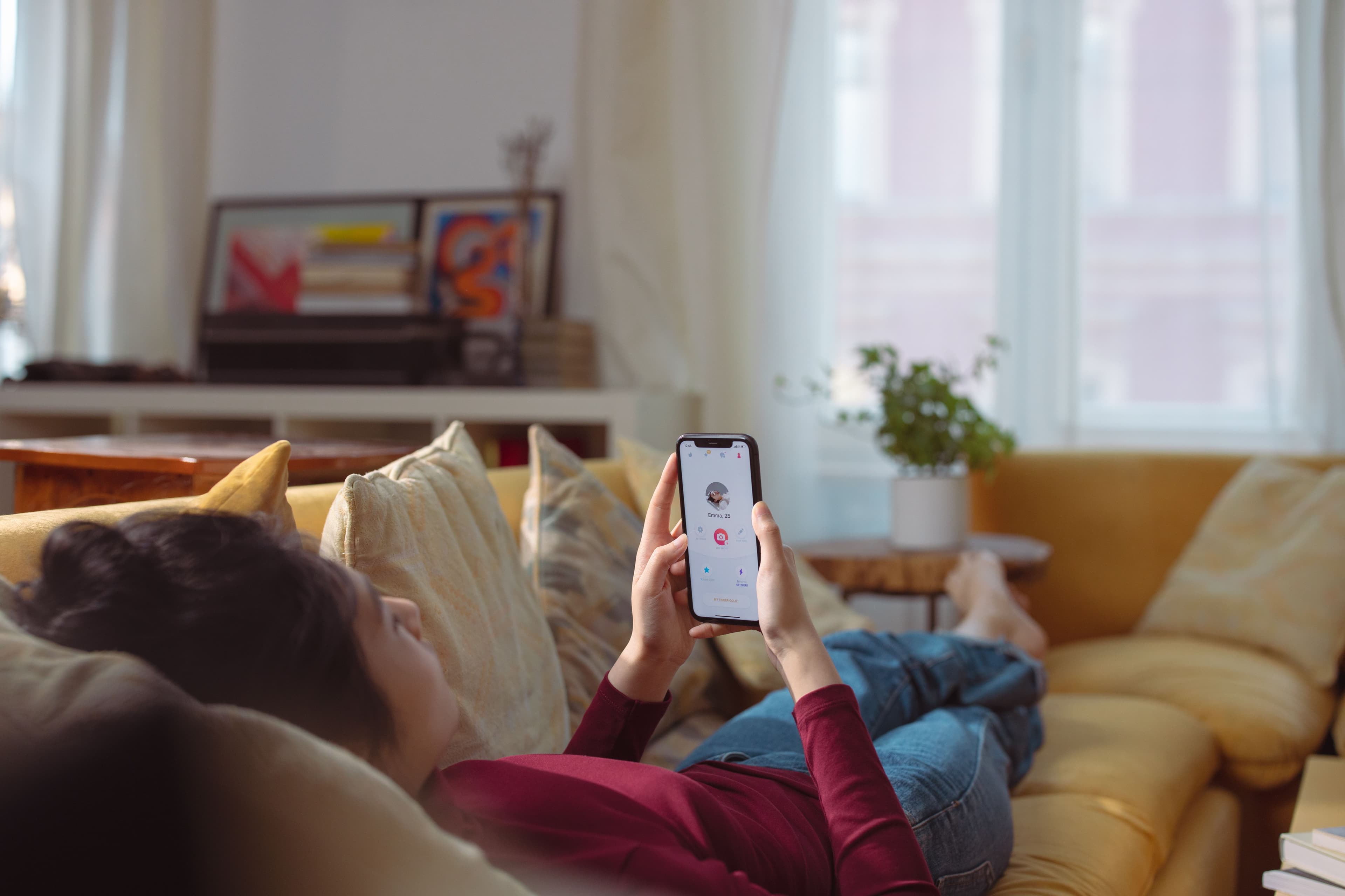 Žmogus, gulintis ant sofos, laiko išmanųjį telefoną su kito žmogaus profiliu ekrane, simbolizuojantis internetinių pažinčių platformą