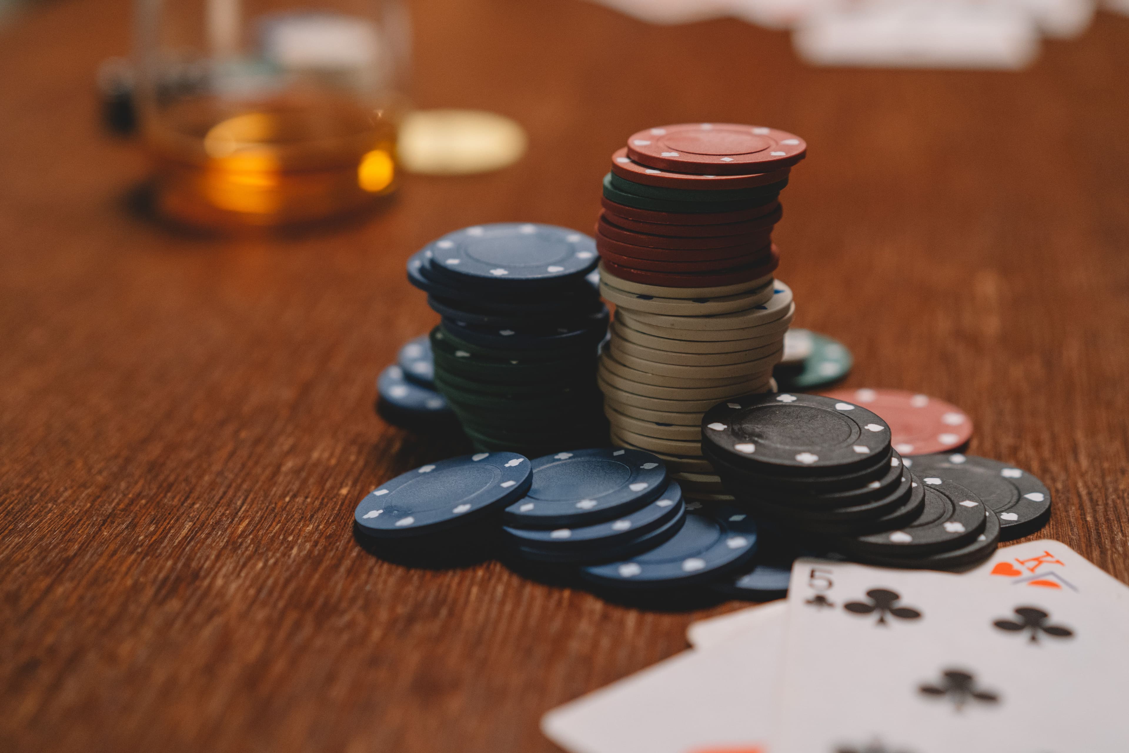 Stacks of blue, red, black and cream poker chips on a wooden table, with playing cards in the foreground and a glass in the background