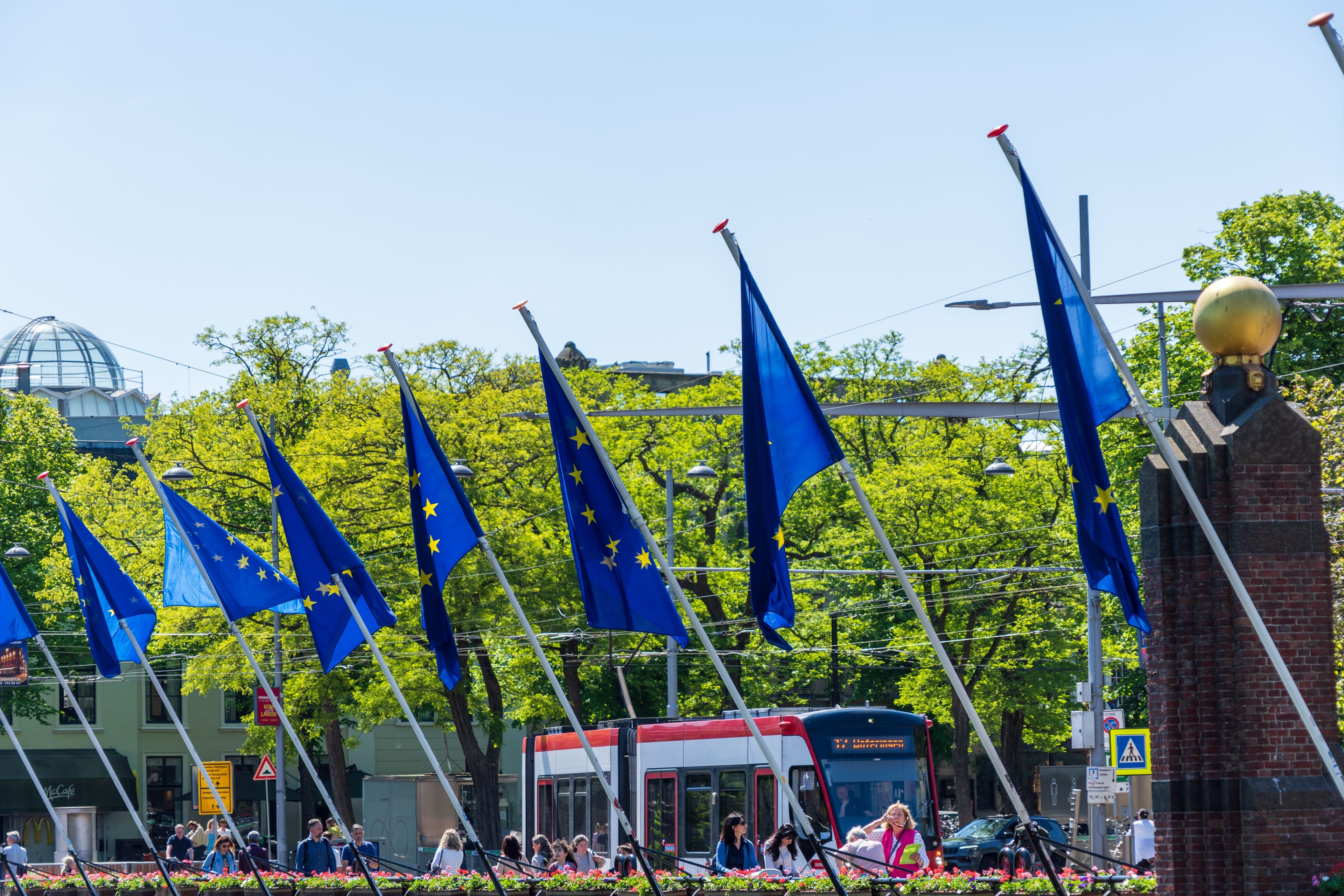 Uma fila de bandeiras da União Europeia em mastros altos ao longo de uma rua soalheira, com um elétrico e árvores verdes ao fundo